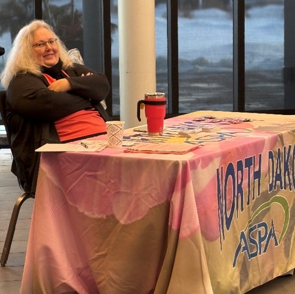 Michelle Buehlmann sitting at a table with the North Dakota ASPA logo on it.